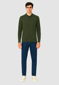 Polo shirt verde a maniche lunghe con un logo discreto, abbinato a jeans blu e sneaker bianche. Tessuto morbido, vestibilità casual.