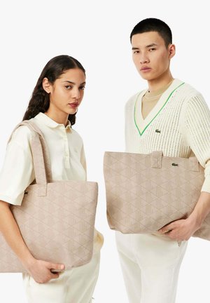 Femme et homme portant chacun des tenues crème clair, tenant des sacs fourre-tout beige avec des motifs géométriques subtils et des logos Lacoste.