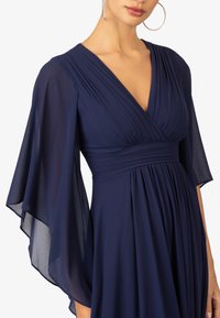 Abito blu navy con scollatura a V profonda, corpetto plissettato e maniche a campana leggere e trasparenti. Tessuto morbido con una caduta delicata.