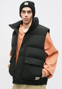 Svart ärmlös puffer väst med hög krage, tryckknappar och framficka, bärs över en orange hoodie, i kombination med en svart mössa.