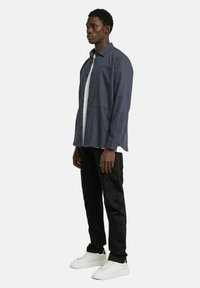 Veste chemise bleu marine avec poche poitrine, portée sur un t-shirt blanc, associée à un pantalon noir et des baskets blanches. Design minimaliste.