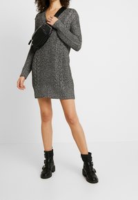 Robe en maille grise avec un motif texturé, manches longues et col en V. Associée à des bottines noires et un sac bandoulière noir.