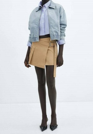 Model iført en lys denimjakke over en blåstribet skjorte, beige plissert mininederdel med sidelås, sorte spidse hæle, stående på hvid baggrund.
