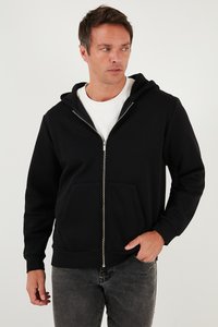 Crna zip-up dukserica s prednjim džepom, napravljena od mekog materijala. Ima kapuljaču koja se može podešavati uzicom i rebraste rukave. Nosiva preko bijele majice.