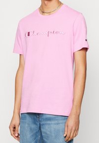 Růžové bavlněné tričko s vyšívaným logem "Champion", kulatým výstřihem, krátkými rukávy a volným střihem. Džíny jsou modré džínoviny.