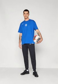 Blaues Nike-T-Shirt mit rundem FC-Logo. Kombination mit schwarzen Hosen in sportlichem Schnitt, hält einen gemusterten weißen und schwarzen Fußball.