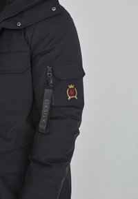 Lähikuva mustasta takin hihasta, jossa on vetoketjulla varustettu tasku, kangasmerkki, jossa lukee "SIKSILK", ja kirjailtu punainen ja kultainen vaakunamerkki.