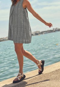 Robe sans manches à rayures grises et blanches avec une coupe décontractée, associée à des sandales à plateforme noires avec des brides blanches. Fond aquatique.