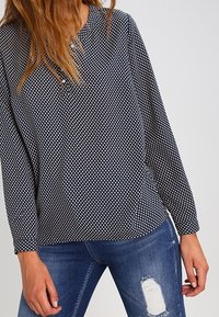 Haut à manches longues bleu marine avec des pois blancs, doté d'un col rond et d'un détail bouton, associé à un jean bleu déchiré.