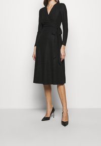 Musta kietaisumekko, jossa pitkät hihat ja V-aukko. Vyötäröllä solmittava nauha, ja helma ylettyy puoliväliin pohjetta. Sileä, teksturoitu kangas.