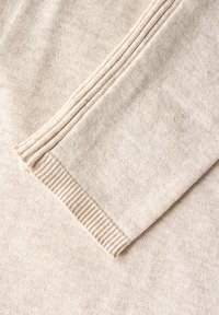 Beige stickad tröja med ribbade ärmslut och textilstruktur, med ett subtilt mönster och mjukt utseende. Fokus på ärmdetalj.