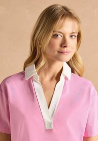 Ružová polokošeľa s krátkym rukávom a bielym golierom, s hlbokým V-výstrihom a jemnou textúrou. Minimalistický dizajn a kontrastné farby.