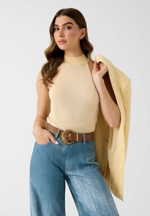 Femme aux longs cheveux bruns portant un haut beige sans manches, un jean bleu à jambe large avec une ceinture marron et tenant une veste jaune sur l'épaule.