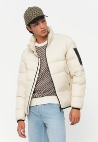 Homme portant une doudoune beige sur un pull à motifs, un jean bleu clair et une casquette à carreaux, regardant sur le côté sur fond blanc.