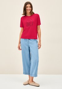 T-shirt rouge à manches courtes avec un design graphique noir circulaire, associé à un jean large bleu clair et des chaussures à enfiler beiges.
