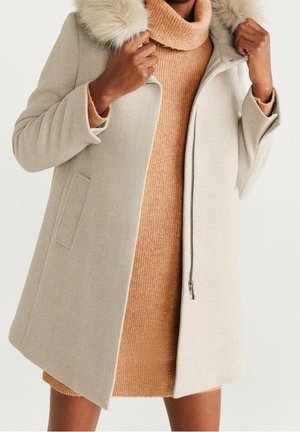Donna che indossa un cappotto di lana beige con collo in pelliccia sopra un vestito a coste color cammello, che sistema il colletto del cappotto con entrambe le mani.