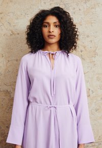 Vestido lilás de manga longa com decote redondo e detalhe de laço. Apresenta uma cintura ajustada e uma silhueta fluida. Tecido suave e macio.