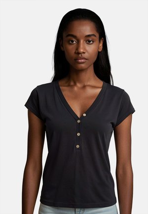 Giovane donna con pelle scura e lunghi capelli neri che indossa una maglietta nera a maniche corte con scollo a V e tre bottoni e jeans azzurri chiari.
