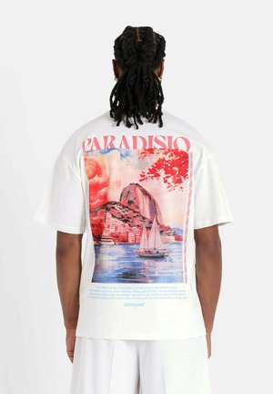 T-shirt blanc à manches courtes avec une impression graphique colorée au dos, représentant une scène côtière avec des voiliers et des accents floraux vibrants.
