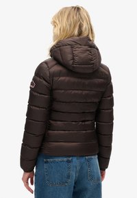 Chaqueta acolchada marrón con capucha, que presenta un diseño de acolchado horizontal, textura suave y un pequeño logo en la parte superior de la espalda.