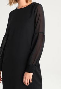 Schwarzes Kleid mit langen, transparenten Ärmeln und gerafften Manschetten, rundem Ausschnitt und einer schlichten, geraden Silhouette. Minimalistisches Design.