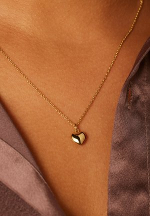 Goldherz-Anhänger an einer zarten Kette mit glänzender Oberfläche, hängend vor einem strukturierten, gebräunten Hauthintergrund.