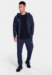 Marine-blauwe ritsjack met capuchon over een zwart T-shirt, gecombineerd met marine-blauwe joggingsbroek. De outfit heeft een gladde textuur en logodetails.