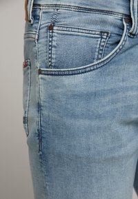 Jeans en denim bleu clair au design ajusté, style classique à cinq poches, avec une légère décoloration sur le tissu pour ajouter de la texture.