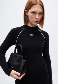 Μαύρο ριμπ πλεκτό πουλόβερ με λευκές ραφές ως διακοσμητικά στοιχεία, με μικρό λογότυπο. Κρατά δομημένη μαύρη τσάντα χειρός με κοντές λαβές.