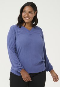 Bluză cu decolteu în V din țesătură violet texturată, cu mâneci lungi și pliate, având o croială lejeră. Purtată cu pantaloni negri.