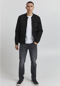 Giacca bomber nera trapuntata con collo rotondo, zip frontale e una tasca sul petto, abbinata a jeans in denim grigi e sneakers nere.