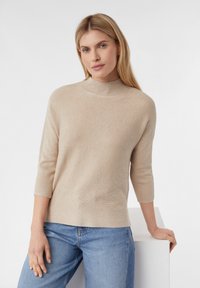 Beige geribbelde gebreide trui met een hoge hals, driekwart mouwen en een ontspannen pasvorm, gecombineerd met blauwe jeans, leunend tegen een witte achtergrond.