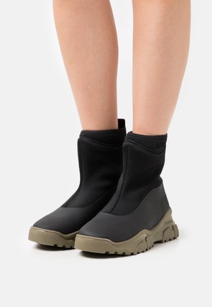 Bottines noires en matériau lisse et extensible avec une semelle en caoutchouc beige épaisse. Présente une tige haute avec un logo discret et une pointe arrondie.