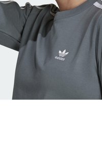 Grå bomullströja med korta ärmar, med rund halsöppning och vit broderad logotyp på bröstet. Utsmyckad med två vita ränder på axlarna.