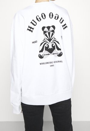 Persona che indossa una felpa bianca con un orsetto astratto nero e la scritta "HUGO," "WORLDWIDE ORIGINAL 1993" sulla schiena.
