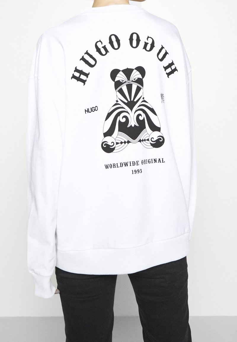 Personne portant un sweat-shirt blanc avec un ours en peluche abstrait noir et le texte "HUGO," "WORLDWIDE ORIGINAL 1993" dans le dos.