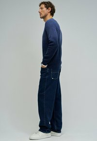 Maglione lavorato a maglia blu navy con texture a coste, abbinato a jeans a gamba larga blu scuro e sneaker bianche; soggetto in posizione di profilo.