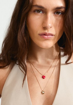 Collane d'oro con due pendenti a cuore; uno rosso e uno in oro metallico, indossate sopra un top beige. Le catene sono sottili con un design delicato.