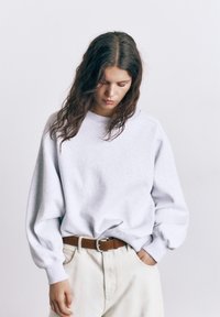 Kvinde med langt, bølget hår, der kigger ned, iført en lysegrå, oversized sweatshirt, beige bukser og et brunt læderbælte.