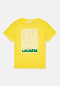 T-shirt en coton jaune avec un graphique carré blanc présentant un motif répété et "LACOSTE" en vert en bas. Manches courtes, col rond.