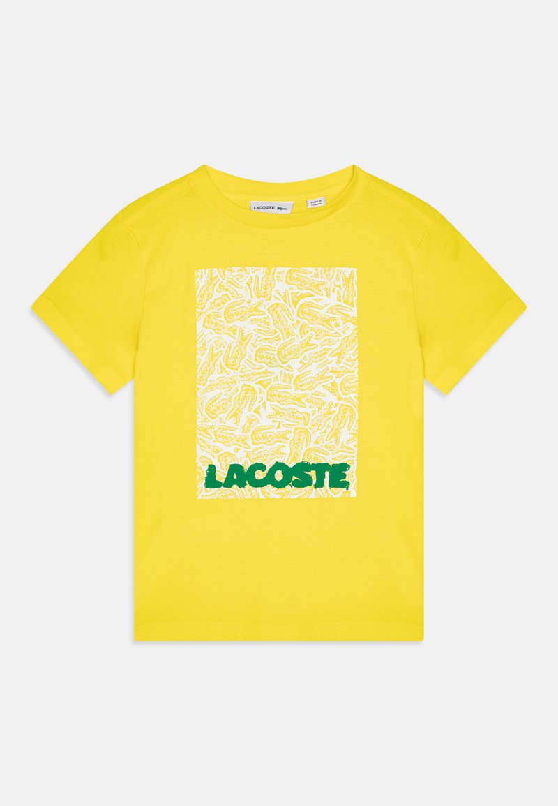 Gul bomull T-skjorte med et hvitt firkantet grafisk mønster med en gjentatt design og "LACOSTE" i grønt nederst. Korte ermer, rund hals.