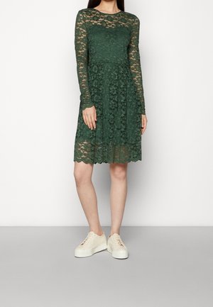 Femme portant une robe mi-longue en dentelle vert foncé à manches longues et des baskets blanches à plateforme, debout devant un fond uni.