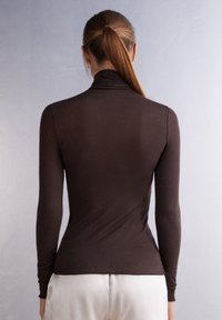 Braunfarbener Langarm-Pullover mit Rollkragen, figurbetont und mit glatter Textur, verfügt über einen hohen Kragen und ein elegantes Design. Weiße Hose teilweise sichtbar.