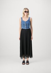 Avskåret denimtopp med knapper, sammen med et langt svart plissert skjørt og svarte åpne sko. Utseendet fullføres med solbriller.