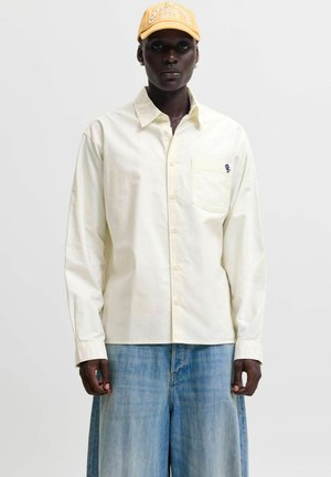 Personne à la peau foncée portant une chemise beige à boutons avec une poche poitrine, un jean ample bleu clair et une casquette jaune avec un texte blanc.