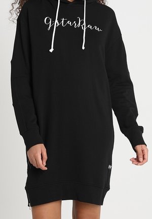 Personne portant une robe sweat à capuche noire à manches longues avec des cordons blancs et un texte cursif blanc sur la poitrine indiquant "gstarkbau."