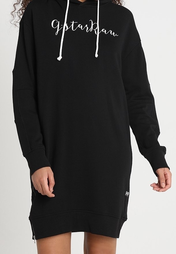 Personne portant une robe sweat à capuche noire à manches longues avec des cordons blancs et un texte cursif blanc sur la poitrine indiquant "gstarkbau."