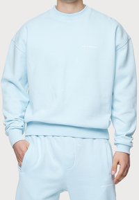 Sudadera azul clara hecha de material suave, con cuello redondo y puños de canalé. Pequeño texto de logo blanco en la parte delantera, diseño minimalista.