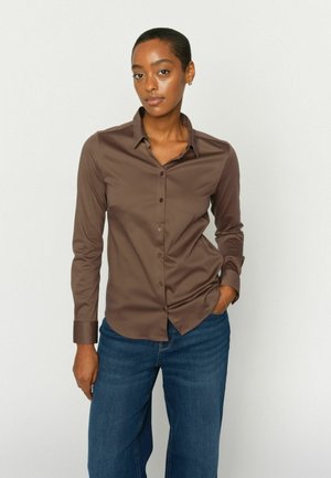 Žena s krátkymi vlasmi, oblečená v hnedej košeli na gombíky a modrých džínsoch, stojaca s jednou rukou v vrecku na jednoliatom pozadí.