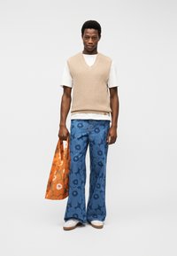 Άνδρας που φοράει μπεζ πλεκτό γιλέκο πάνω από λευκό T-shirt, μπλε τζιν παντελόνι με φαρδιά πόδια και φλοράλ μοτίβο, λευκά αθλητικά παπούτσια, κρατώντας πορτοκαλί φλοράλ τσάντα ώμου, όρθιος.